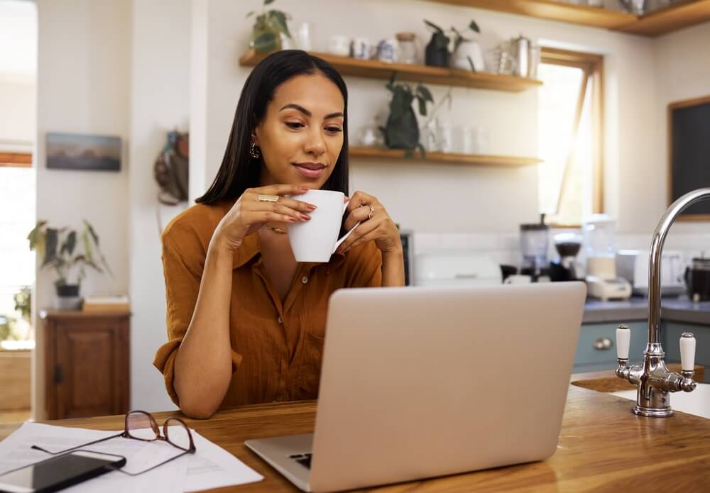 Mulher de cabelo preto sentada em frente a um notebook, enquanto segura uma xícara. Ela está refletindo e observando a tela.