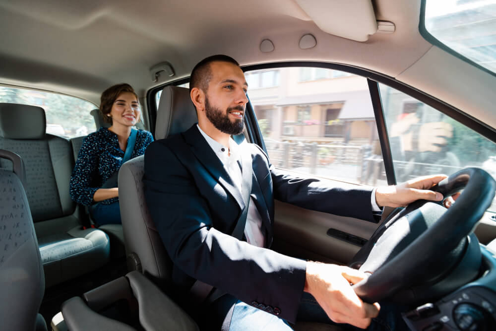 Imagem de um homem dentro de um carro. Ele está dirigindo, pois trabalha com serviços de transporte como fonte de renda extra. Ao fundo é possível ver uma passageira, uma mulher.